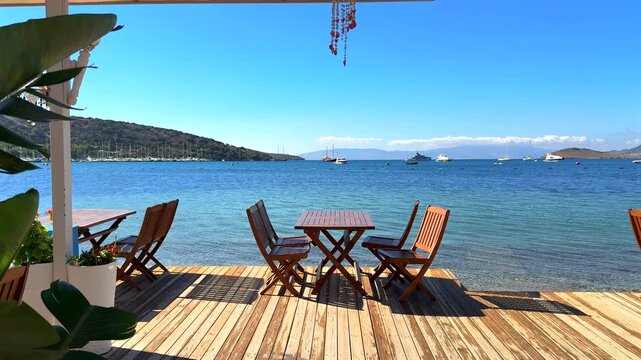 Seaside dining deck with wooden tables and chairs overlooking a beautiful turquoise bay, sailboats, and distant hills under a clear blue sky.