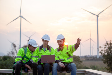 Three engineer in safety uniforms and helmets sitting on platform with wind turbines at sunset, discussing clean energy project. Concept of renewable energy, teamwork, sustainability and wind power.