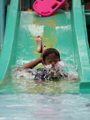 A young boy is joyfully sliding down a green water slide face-first, with water splashing over his face, enjoying a thrilling ride at the outdoor water park.