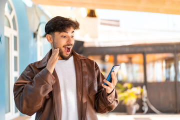 Handsome Arab man at outdoors looking at the camera while using the mobile with surprised expression