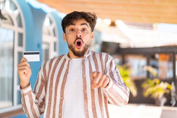 Handsome Arab man holding a credit card at outdoors surprised and pointing front