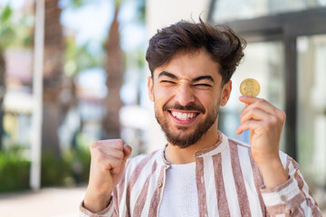 Handsome Arab man at outdoors holding a Bitcoin and celebrating a victory