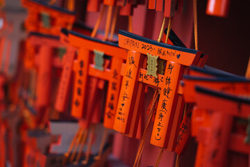 This image features several miniature orange torii gates, typical of Japanese Shinto shrines, hanging in rows. Each gate has black Japanese calligraphy written on it.