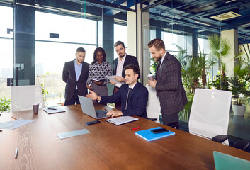 Leader at office desk explaining work task, discuss project, present new creative idea or plan, positive coach gives seminar to multinational diverse team of young employees, teambuilding, mentoring