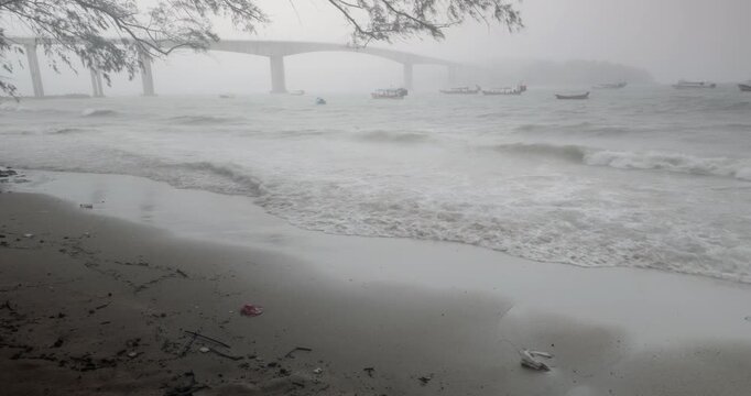 Enveloped in thick, stormy fog, a large bridge and several fishing boats are tossed on the choppy sea while waves continuously wash up on the shoreline.
