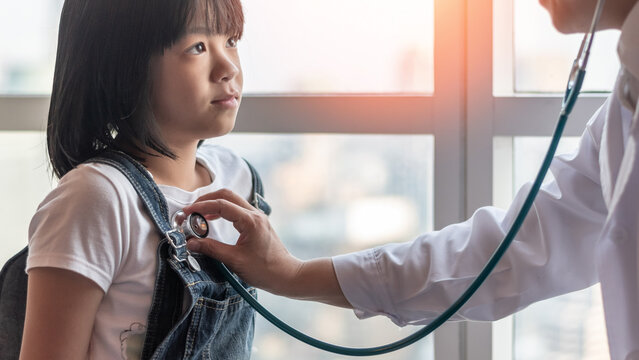Congenital heart disease defect and pediatric care concept. Asian school child patient with pediatrician doctor examining girl kid health checkup with stethoscope in medical clinic