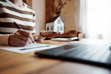 Person signing a real estate contract while holding a small house model, representing home ownership, property investment, and mortgage agreement.