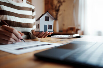Person signing a real estate contract while holding a small house model, representing home ownership, property investment, and mortgage agreement.