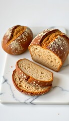 Two loaves of artisan bread, one whole and one sliced, on a white marble cutting board, studio shot.