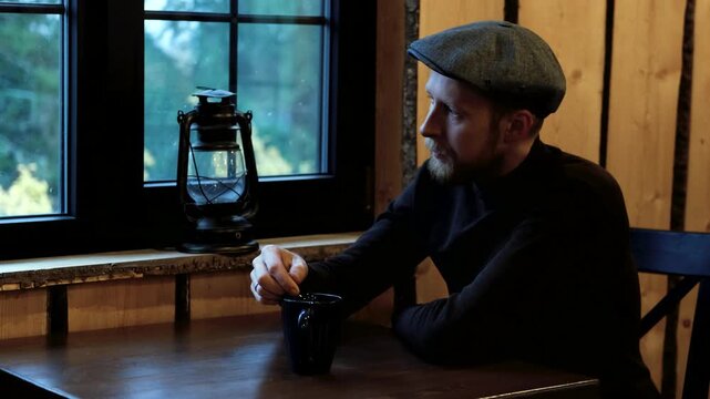 Man in flat cap sits at wooden table, stirring coffee in black mug, with lantern and window in background, camera pans to capture thoughtful ambiance