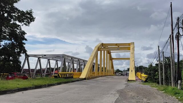 Barru, April 11, 2024 - A yellow steel bridge stands over a road under a cloudy sky, connecting two areas with visible cars, power lines, and nearby houses in an urban setting.