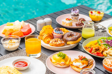 Assorted breakfast dishes by poolside table, featuring pastries, fruits, omelet, beverages. Tropical, vibrant setting, ideal for vacation, summer, travel, lifestyle content.