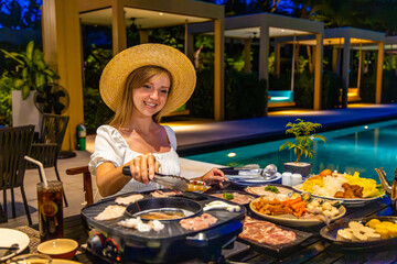 Woman enjoying hot pot dinner poolside at night in outdoor restaurant. Relaxed dining experience with fresh ingredients, vibrant setting, leisure, tourism, culinary adventure.