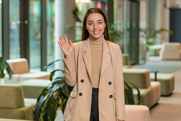 Hello. Happy young Caucasian businesswoman employee waves hand hi gesture greeting welcomes someone, online webinar, remote distance conversation. Entrepreneur woman in formal standing in office lobby © Andrii Iemelianenko