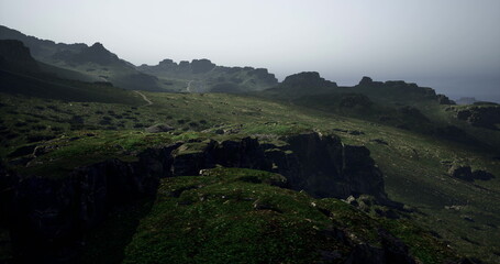 Rolling green hills stretch across the landscape under a soft mist, revealing rugged cliffs and distant rock formations. A peaceful atmosphere envelops the area near the coastline.