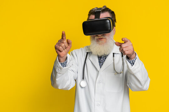 Excited happy senior doctor cardiologist man using headset helmet app, watching virtual reality 3D 360 video. Old apothecary grandfather in VR goggles on yellow background. Future medicine technology
