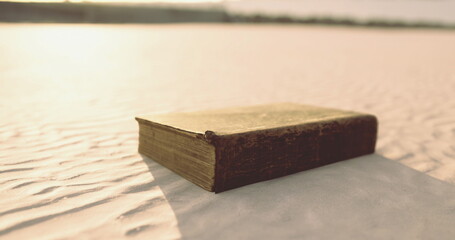 A dusty old book lies on the warm sand under the gentle glow of the sun. The serene coastal landscape creates a peaceful atmosphere, inviting curiosity about the books secrets.