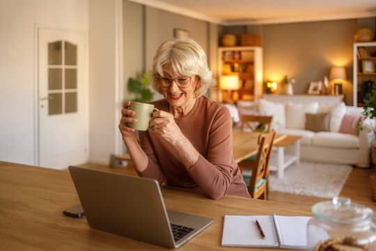 Cheerful senior woman holding coffee cup and connecting with family over video on laptop while relaxing at home
