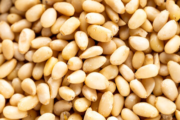 A close-up, full-frame shot of a pile of raw, shelled pine nuts. The light-colored, elongated kernels are scattered, creating a textured background