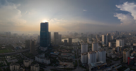 Aerial panorama of Hanoi city skyline at sunset, featuring tall buildings and a calm sky with sun rays breaking through clouds.
