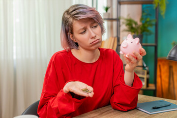 Young woman unhappily counting coins from piggy bank sitting at home table, sighing disappointed. Caucasian girl demonstrating financial shortage and frustration, concerned about insufficient savings.