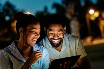 Portrait of young happy couple using tablet computer at night, using modern technology having fun with glowing screen in dark outdoors in the city
