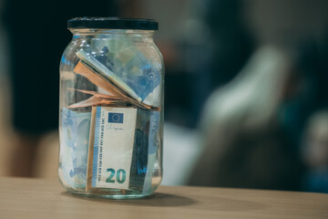 A glass jar filled with euro banknotes sits on a table, symbolizing donations, fundraising, or savings.