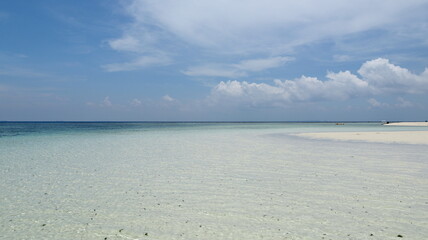 Turquoise water gently laps against the white sand beach under a bright blue sky with fluffy...