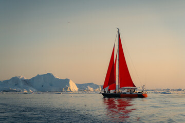 Fototapeta premium A solitary red sailboat courageously navigates through the surreal, glacial landscape of Greenland's Ilulissat Icefjord