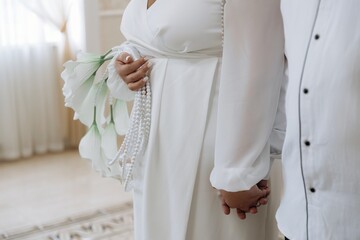 Bride and groom standing together holding hands. Bride wears a white dress and pearls, holding modern white wedding flowers. Intimate ceremony moment