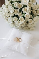 Pair of classic gold wedding bands tied with a white ribbon on a pearl-embellished ring pillow. Bokeh background of white roses