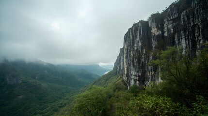 Majestic sheer cliff face covered in lush green vegetation with a misty valley and distant river below under a cloudy sky creating a dramatic natural landscape