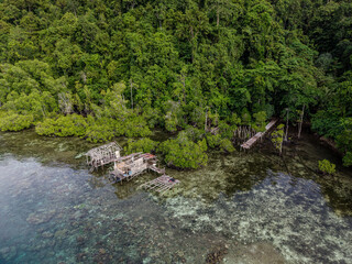 Aerial view of wooden stilt bungalows over clear shallow turquoise water near a tropical island in Raja Ampat, Indonesia.  

