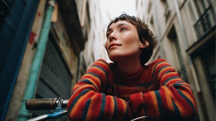 Young person wearing a colorful sweater resting on a bicycle while looking thoughtfully in a narrow urban alleyway during the day