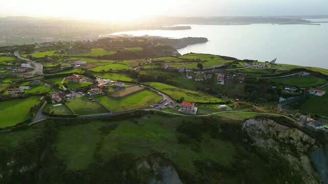 Aerial view of the stunning Asturian coastline at golden hour, featuring lush green fields, charming houses, and the tranquil Bay of Gij?n under a warm sunset glow.