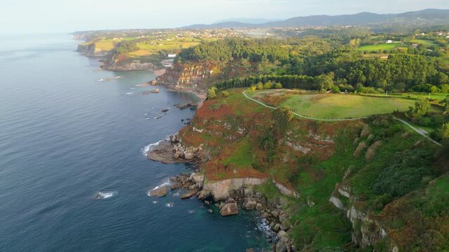 Golden hour illuminates the rugged Asturian coastline, revealing dramatic cliffs, hidden coves, and lush forests. A scenic path winds along the clifftops.
