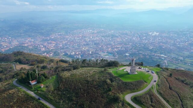 Panoramic aerial view of Oviedo city and the surrounding Asturian landscape, featuring the iconic Cristo del Monte Naranco statue under a cloudy autumn sky.