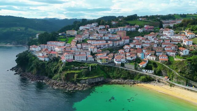 Aerial view of the picturesque fishing village of Lastres, Asturias, Spain. Traditional houses cling to cliffs overlooking a serene beach and bustling marina. A vibrant coastal scene.