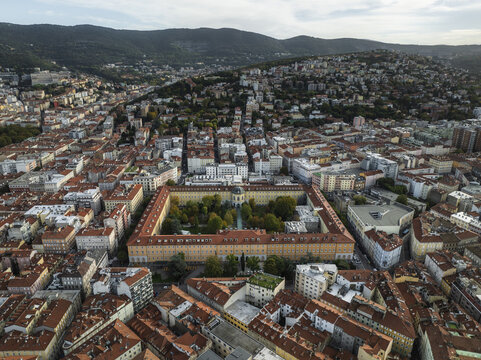 Aerial view of a grand, quadrangular building with a verdant courtyard nestled amidst the terracotta rooftops of the city, embraced by distant, rolling hills, Trieste, Friuli-Venezia Giulia, Italy.
