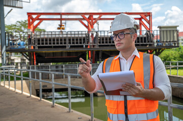 an engineer in safety vest inspects water gate structure, recording data on a sunny day