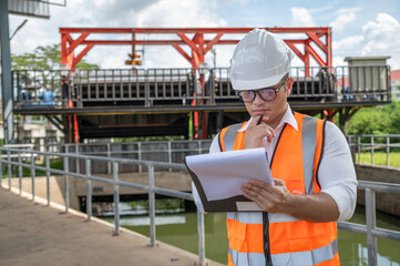 an engineer in safety vest inspects water gate structure, recording data on a sunny day