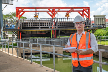 an engineer in safety vest inspects water gate structure, recording data on a sunny day
