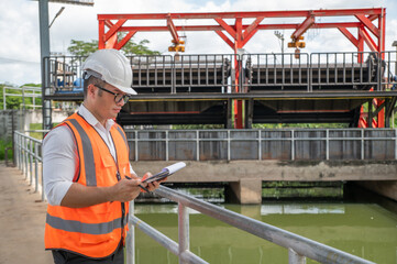 an engineer in safety vest inspects water gate structure, recording data on a sunny day
