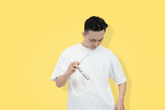 A young Asian man in white shirt holding eyeglasses and looking down with a contemplative expression against a bright yellow background