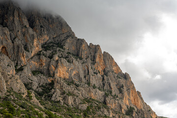 Steep rugged cliffs of Puig Campana mountain in Spain rise into low clouds, showcasing dramatic rock formations and natural textures under a moody, overcast sky.