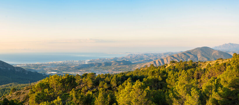 Expansive landscape of Puig Campana region in Spain, featuring lush green pine forests, rolling hills, and distant Mediterranean Sea under a clear sky. Ideal for travel, nature, and tourism.