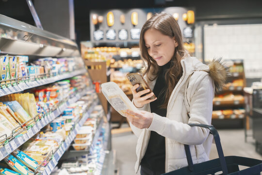 Woman examines a package of cheese in the grocery store, scanning the label with her phone to learn about its ingredients and expiration date while shopping.