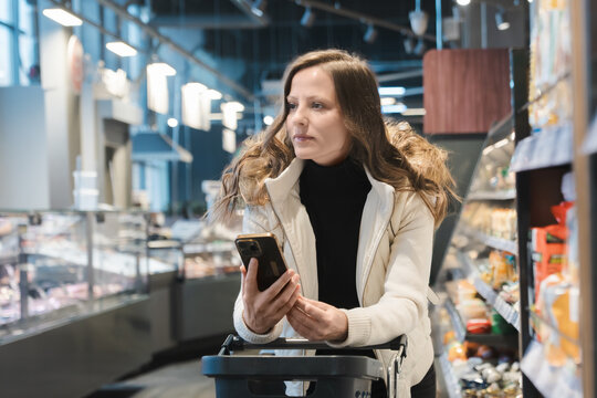 In a modern supermarket, a woman pushes a grocery cart while looking at her smartphone, recalling her shopping plan and list of items to buy amidst various products.