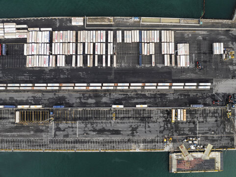Trieste, Italy - 10 October 2025: Aerial view of the bustling port, where rows of containers create a geometric pattern against the dark asphalt, framed by the shimmering turquoise sea.