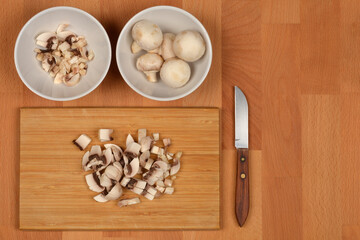 An overhead shot of fresh whole and chopped white button mushrooms in white bowls and on a wooden cutting board with a knife